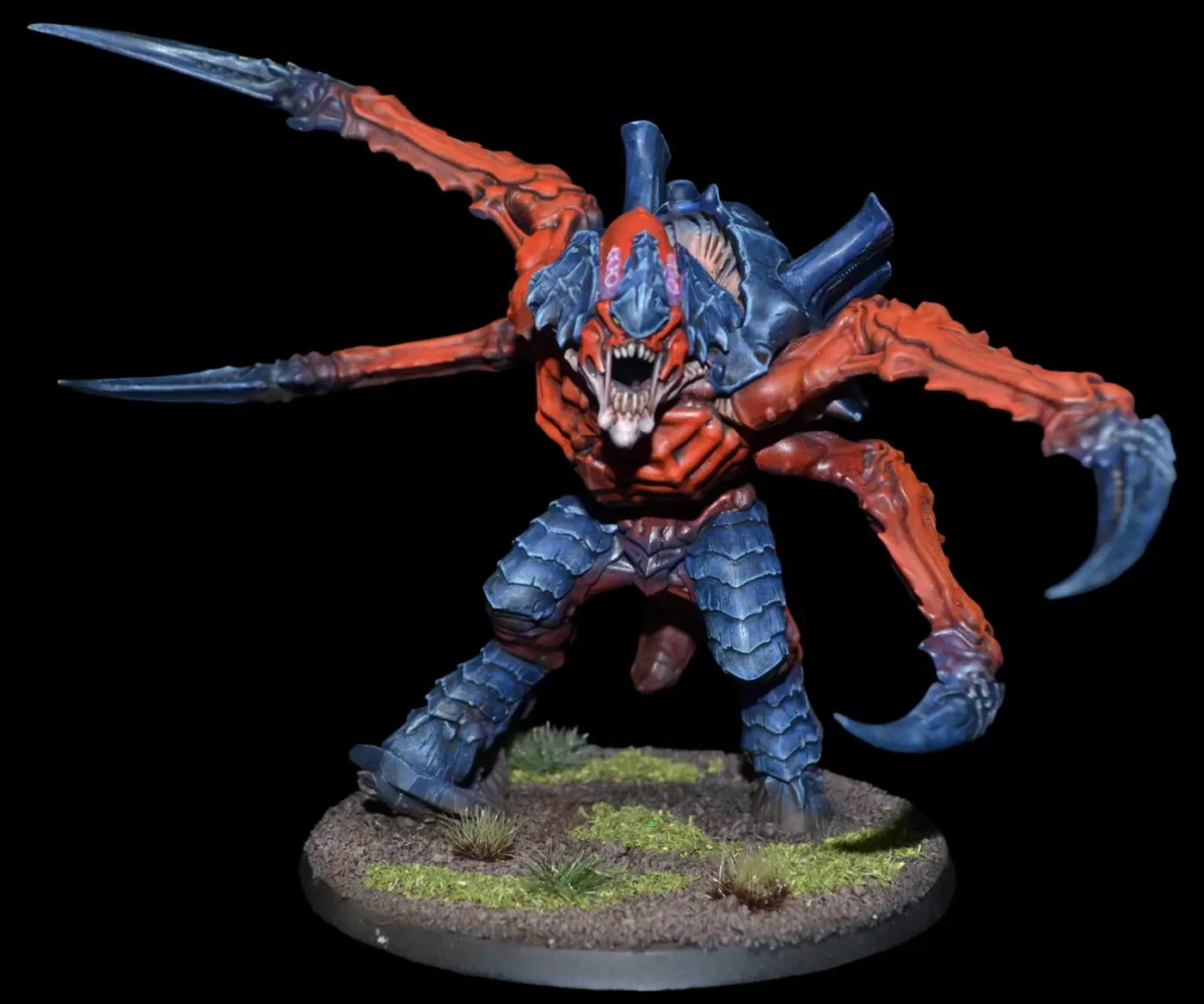 Carnifex mit rotem Körper, blauem Chitinpanzer und ausladenden Klauen auf begrünter Base